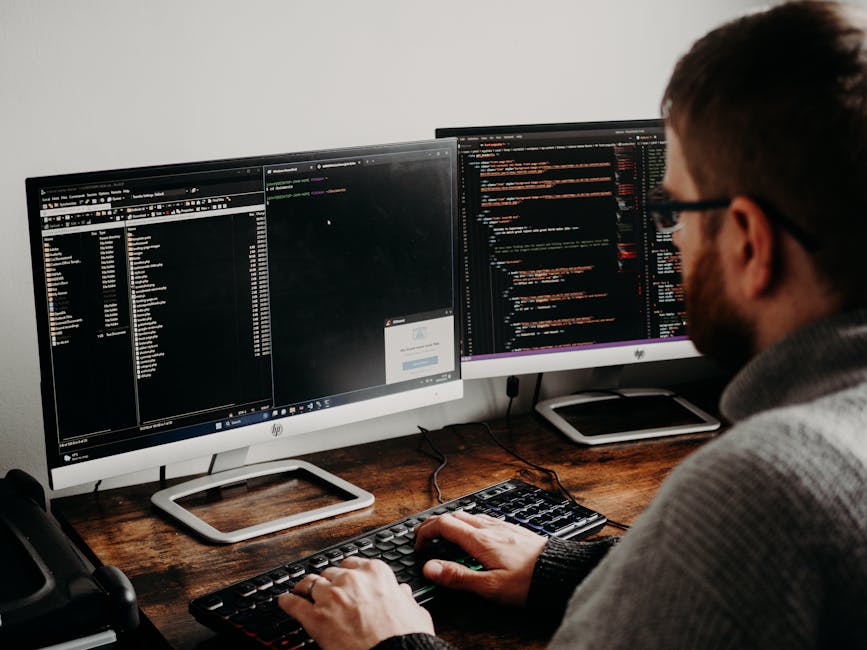 A person with glasses and a beard is seated at a wooden desk working on a computer with dual monitors, both displaying lines of code. The left monitor shows a file directory and code editing interface
