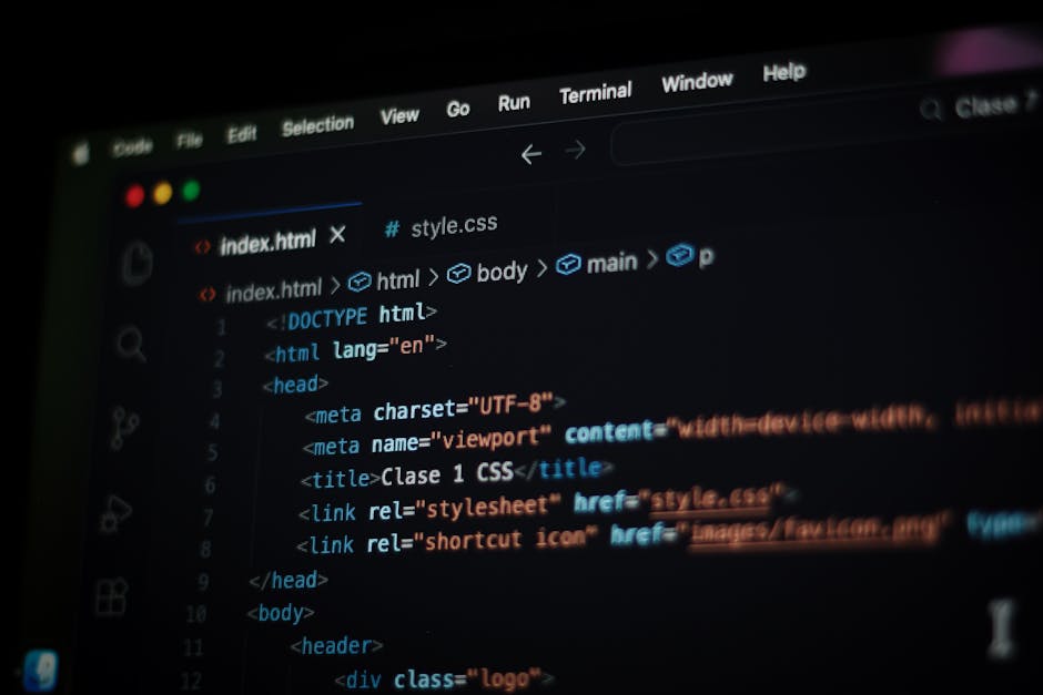 A person with glasses and a beard is seated at a wooden desk working on a computer with dual monitors, both displaying lines of code. The left monitor shows a file directory and code editing interface